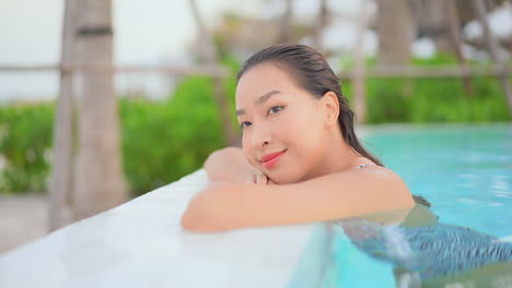 pretty young lady leaning on the edge of a swimming pool with her face and arms, standing half submerged in the water and closing her eyes smiling