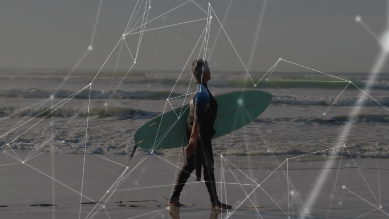 Senior female surfer walking along beach carrying teal surfboard with technology network overlay