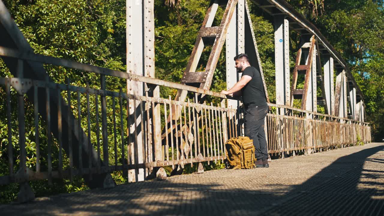 joven viajero con mochila admirando la naturaleza desde un viejo puente oxidado al aire libre en un soleado día de verano