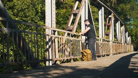 Young-man-traveler-with-backpack-admiring-the-nature-from-a-rusty-old-bridge-outdoors-on-sunny-summer-day
