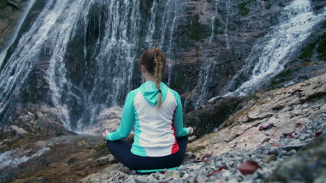 woman in a lotus yoga pose with raised arms overhead, by a waterfall, rear view