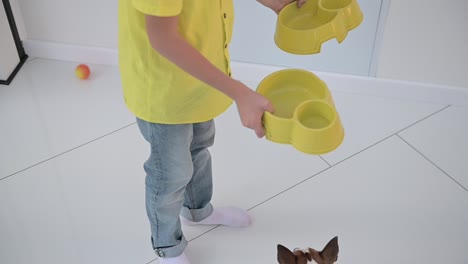 a boy carries two dog bowls, and one of his dogs follows him as he leaves the room