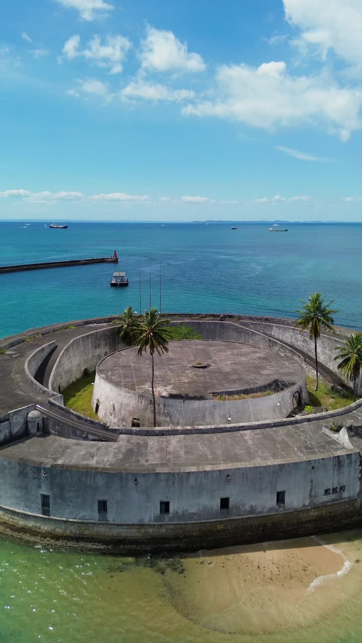 Vertical: Drone flies over São Marcelo Fort, blue water, and anchored boats on a sunny day in Salvador, Bahia, Brazil