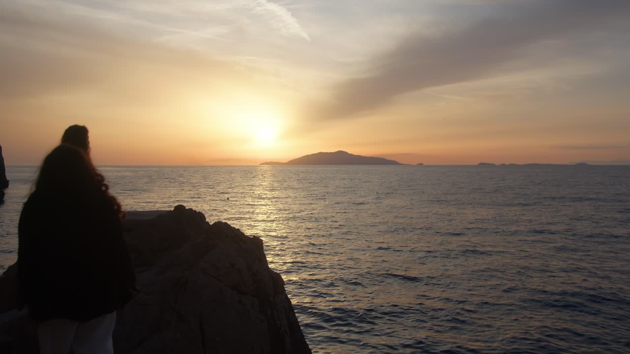 Silhouetted sisters walk toward the rocks to admire the sunset over Capri Island, Italy