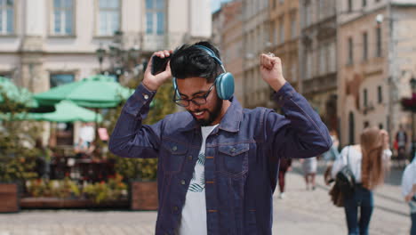 happy young indian man in wireless headphones, listening favorite energetic music in city street