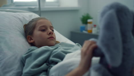 Upset-girl-lying-hospital-bed-portrait.-Sad-kid-playing-with-plush-toy-alone.