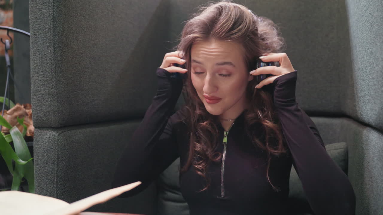 Light skin lady with long curly blond hair wearing black and blue headset around neck smiling while seated comfortably in cozy booth with dark upholstery and plants in background