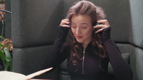 light skin lady with long curly blond hair wearing black and blue headset around neck smiling while seated comfortably in cozy booth with dark upholstery and plants in background