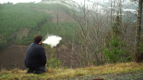 un joven milenario arrodillado y usando una máquina de vape y soplando humo mientras disfruta de estar al aire libre en la naturaleza, en cámara lenta