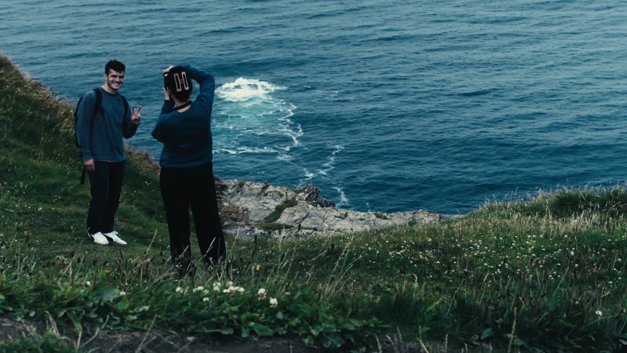 Overhead view of a smiling man posing as a woman takes his photo, with flowers, greenery and waves in background - Ireland