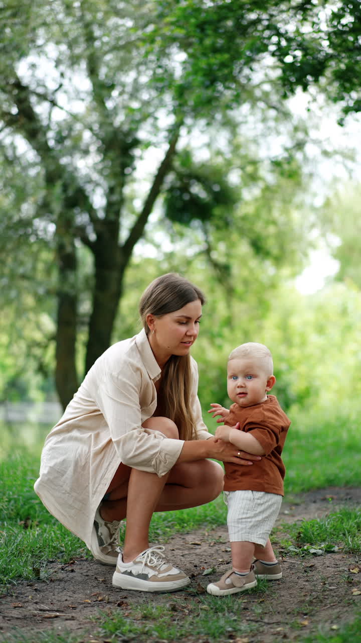 Mother sits squatted near her little son. Woman holds her lovely baby standing unsteadily. Vertical video.