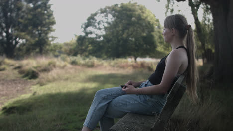 a woman sitting in the bench under the shade of tree and looking out at the distance