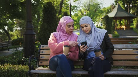 two muslim females in casual clothes and hijabs. they smiling, enjoying coffee and looking at screen of smartphone. sitting on bench in park. сlose up