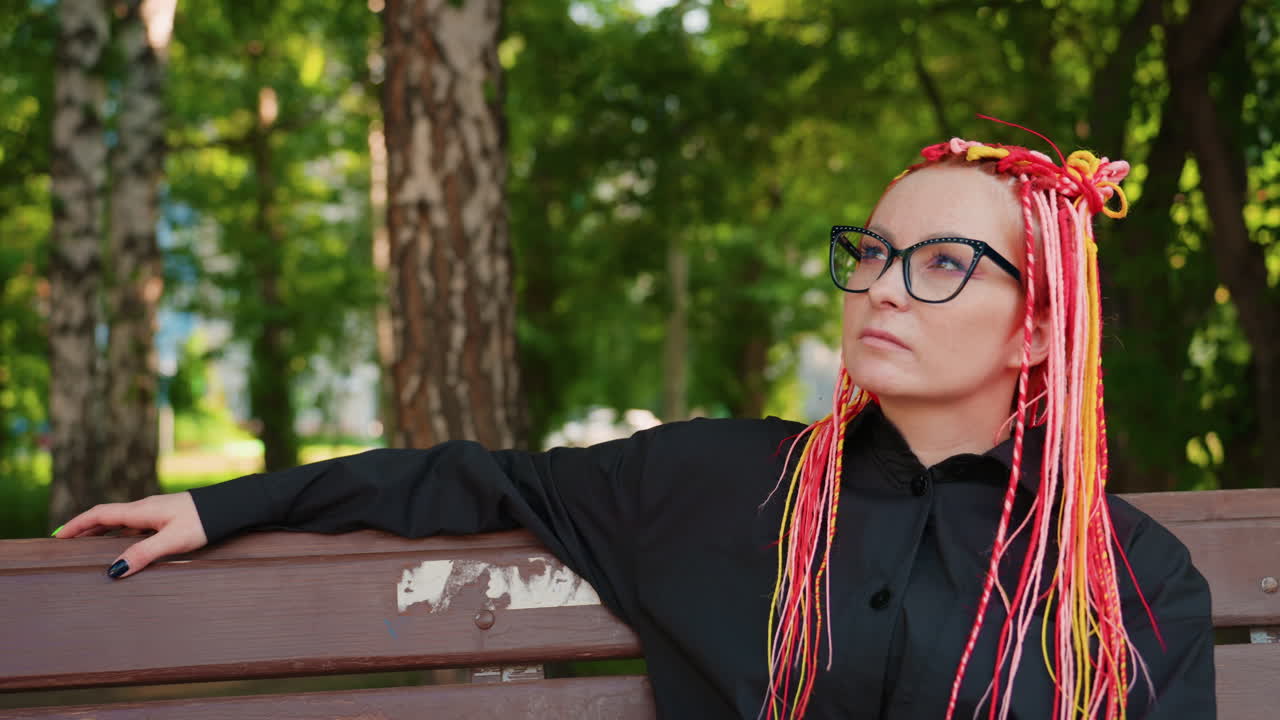Mujer caucásica con trenzas rosas sentada en un banco de madera en un parque frondoso, mirando hacia arriba de forma contemplativa, con gafas y chaqueta negra, árboles de abedul iluminados por el sol de fondo, ambiente de poeta introspectivo