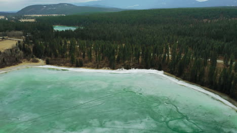 Frozen-teal-green-lake-with-woods-and-mountains-in-the-distance