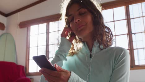 mujer de raza mixta feliz preparándose para el entrenamiento, sosteniendo un teléfono inteligente poniendo auriculares en una habitación soleada