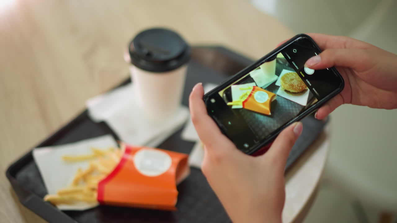 mujer capturando una foto de su comida usando su teléfono inteligente, centrándose en el arreglo de comida de papas fritas, una hamburguesa y una taza de café en una bandeja negra, la imagen se muestra en la pantalla del teléfono