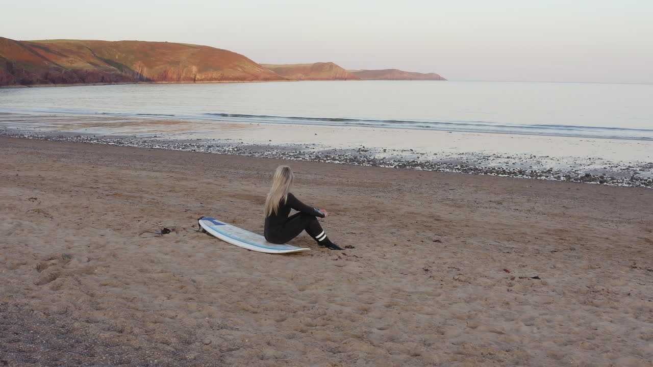 서프보드 에 앉아 바다 를 바라보는 웨이트 슈트 를 입은 여자 의 드론 촬영