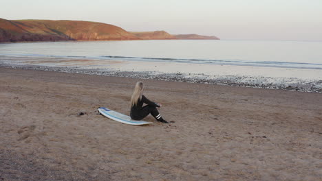 drone shot of woman wearing wetsuit sitting on surfboard and looking out to sea