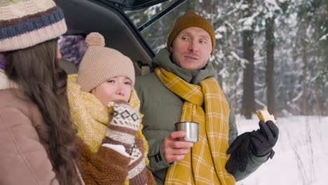 side view of parents and daughter eating sandwiches and drinking hot drink sitting in the trunk of the car in a snowy forest 1