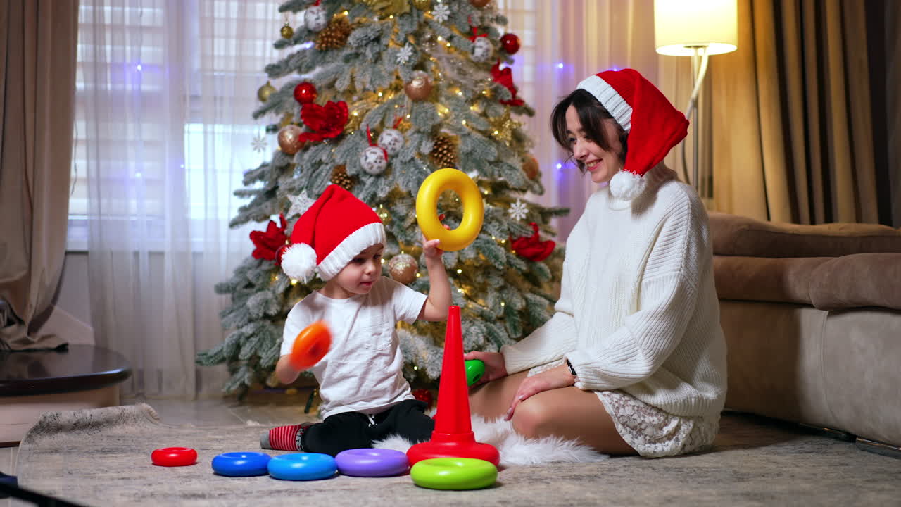 Mother and son wearing red Santa caps spend a cozy evening at home near the Christmas tree. Lovely baby boy constructs a toy pyramid.
