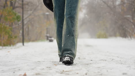 low angle view of someone in black boots and jeans, carrying an item behind them while walking on a snowy path during snowfall, with trees and a bench in the serene winter landscape