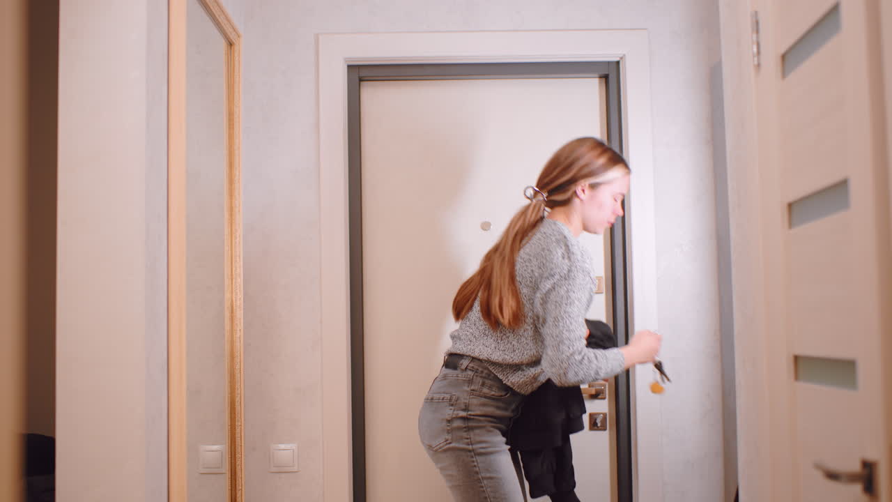 Confident young woman standing in hallway holding black jacket then go out, facing front door with calm focused expression, casual modern outfit