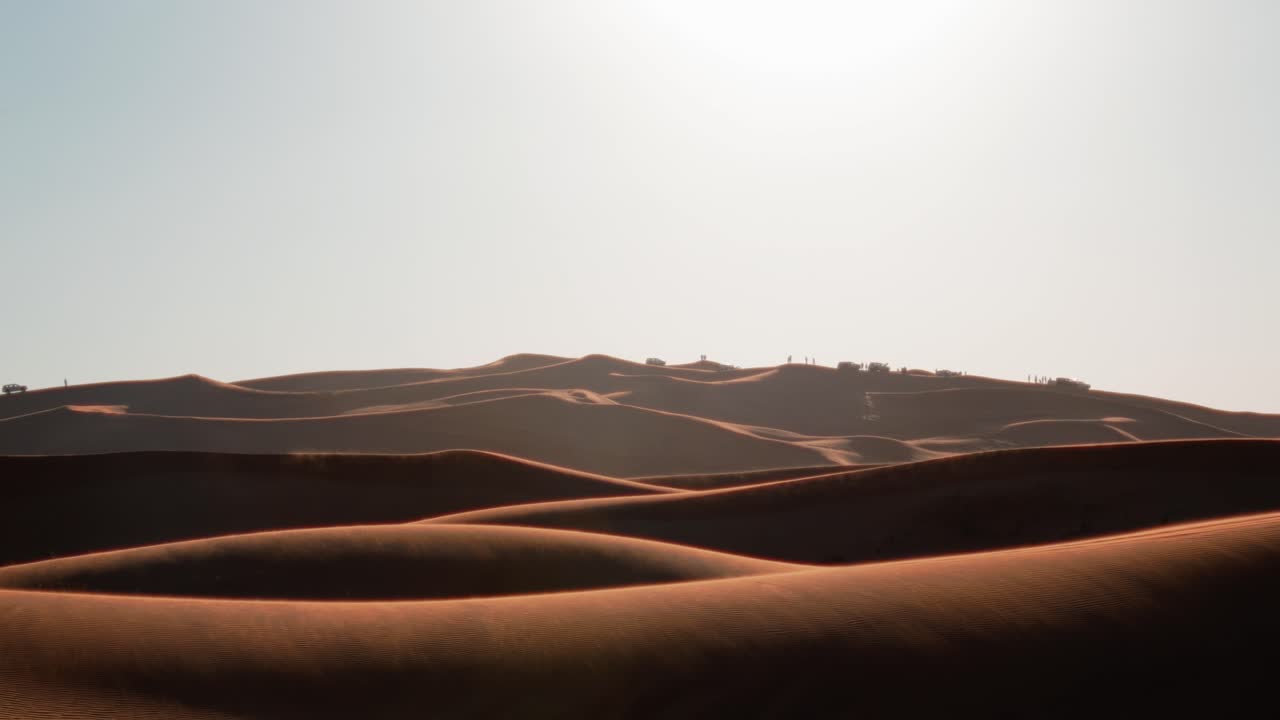 La luce solare intensa si proietta su dune di sabbia ondulate, sfondo naturale ripreso a mano con leggero tremolio, la sabbia si solleva dalla cima a causa del vento