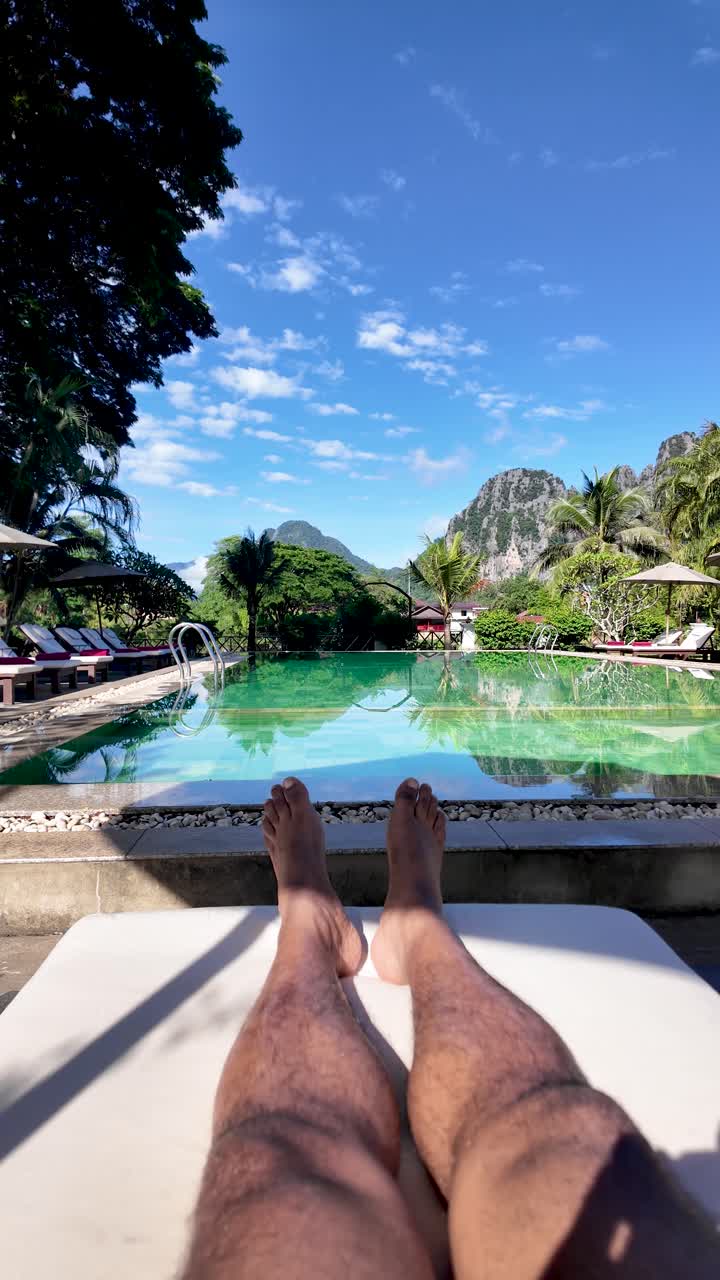Person's legs relaxing on a sun lounger by the swimming pool at a resort with mountains. vertical video