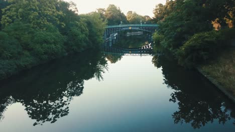 Drohnenluftfluss-In-Einem-Park-Bei-Sonnenaufgang-Mit-Einer-Brücke,-Die-Auf-Dem-Wasser-Reflektiert