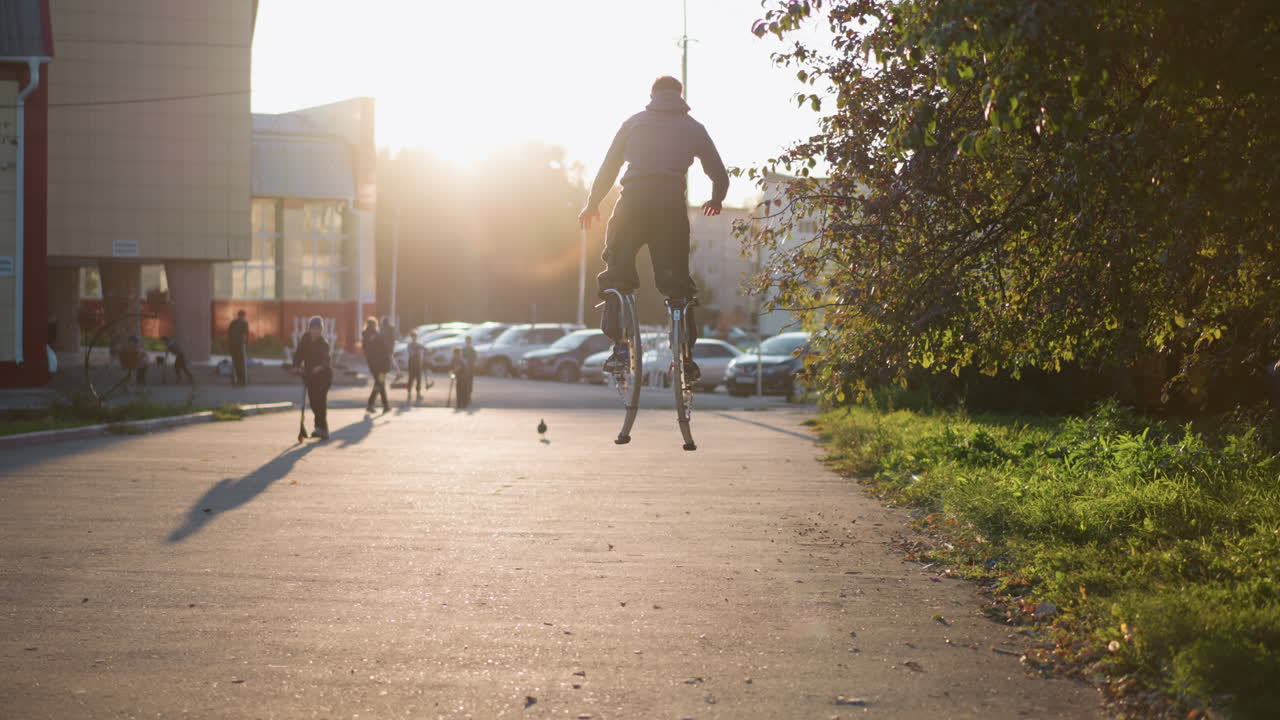 회색 후디와 어두운 회색 바지를 입은 사람이 집으로 둘러싸인 주거 지역에서 해가 지는 동안 다리에 스으로 뛰어내리는 뒤쪽 사진, 도시 에너지, 창의성 및 역동적인 움직임을 보여줍니다.