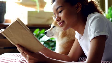 fotografía de cerca de una chica de raza mixta dueña de un perro leyendo un libro acostada en la cama en un apartamento moderno cerca de una adorable mascota obediente. interesante pasatiempo, literatura y concepto de animales.