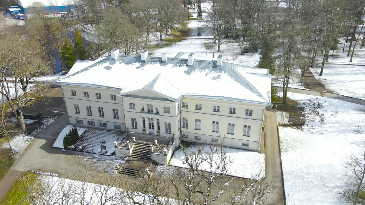 Aerial high angle view of large preserved manor in Saku, Estonia. White mansion house surrounded by trees and pathways in park. Green lawn covered with snow. Fancy neoclassical building and facade.