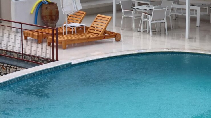 Rain Drops Falling Onto Water In Swimming Pool On Rainy Holiday In Greece With Wet Sunbeds