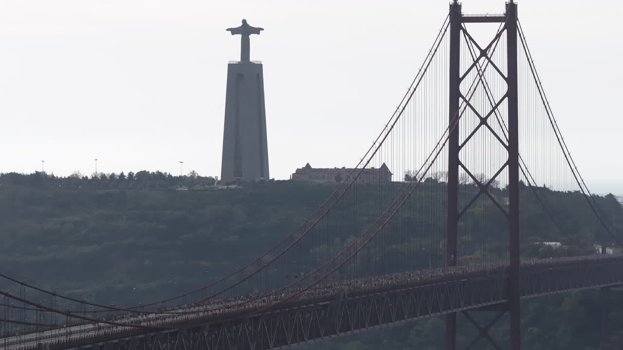 Cinematic aerial drone view of thousands of runners crossing the 25th April suspension bridge in Lisbon, Portugal, with the Cristo Rei monument overlooking the race