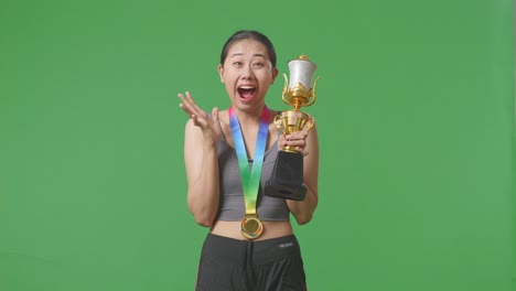 asian woman with a gold medal looking at a gold trophy in her hands and saying wow being happy winning as the first winner on green screen background in the studio