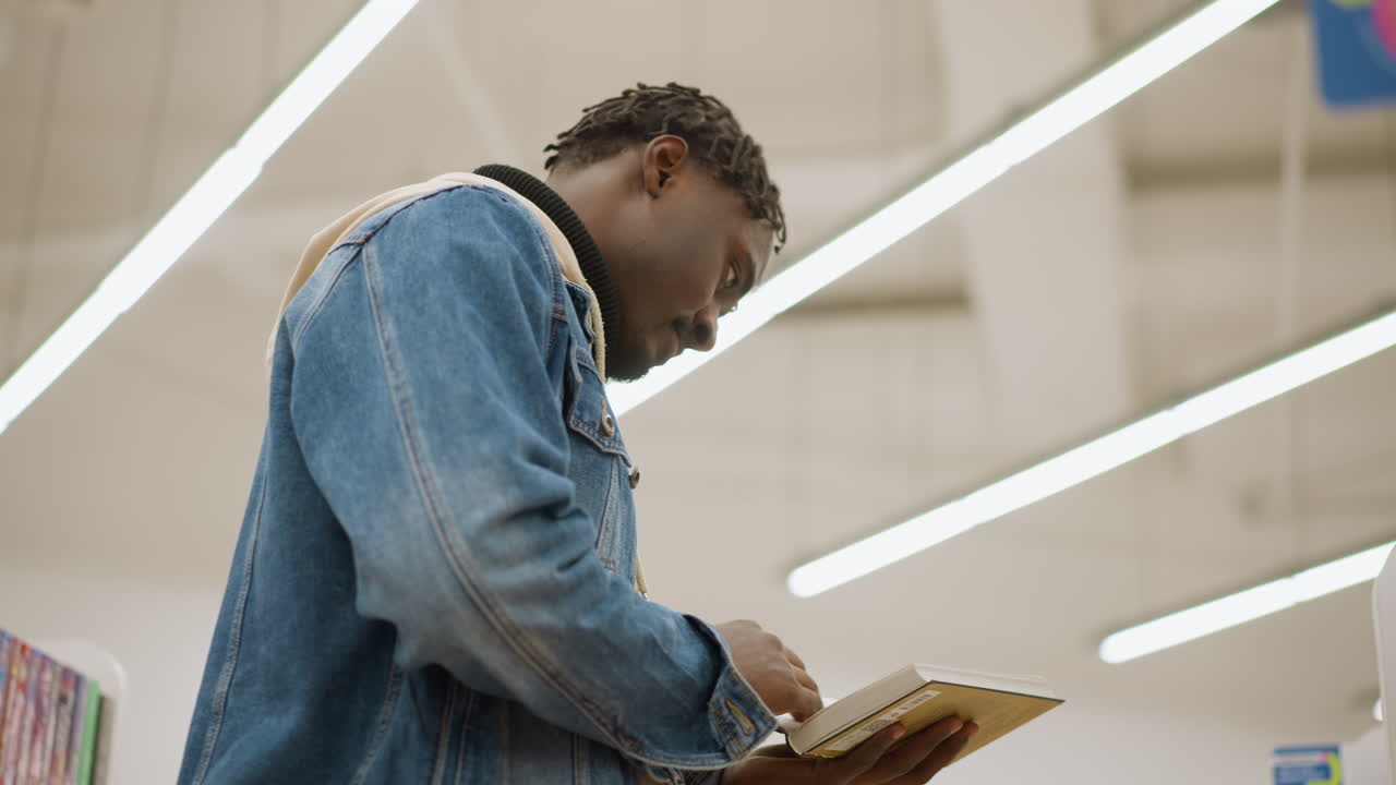 Close-up shot of man in denim jacket reading book in library, focused on text under bright overhead lights, browsing through pages with calm demeanor in modern environment