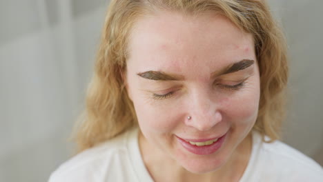 close up of beautician using wooden stick to apply wax on client eyebrow during grooming treatment and client smiles gently with eyes closed while beautician wearing black gloves performs application