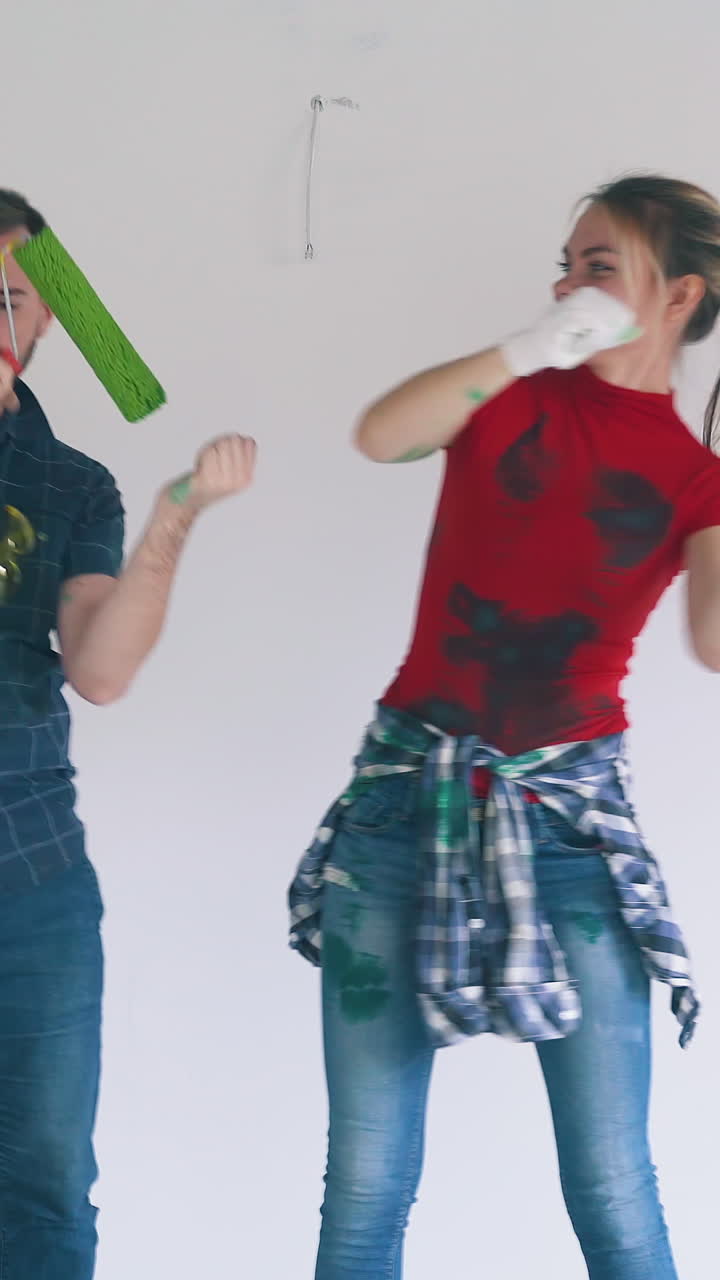 Dolly cam shot: joyful guy and long haired girl with painting tools dance near white wall with stains in empty spacious room