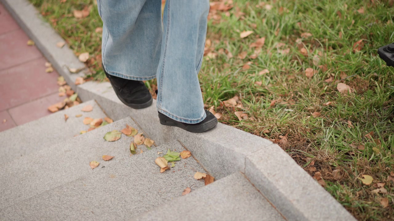 Close up of person legs and feet as they descend stone steps wearing light blue jeans and black shoes. scene includes scattered autumn leaves on grassy area beside red brick pavement