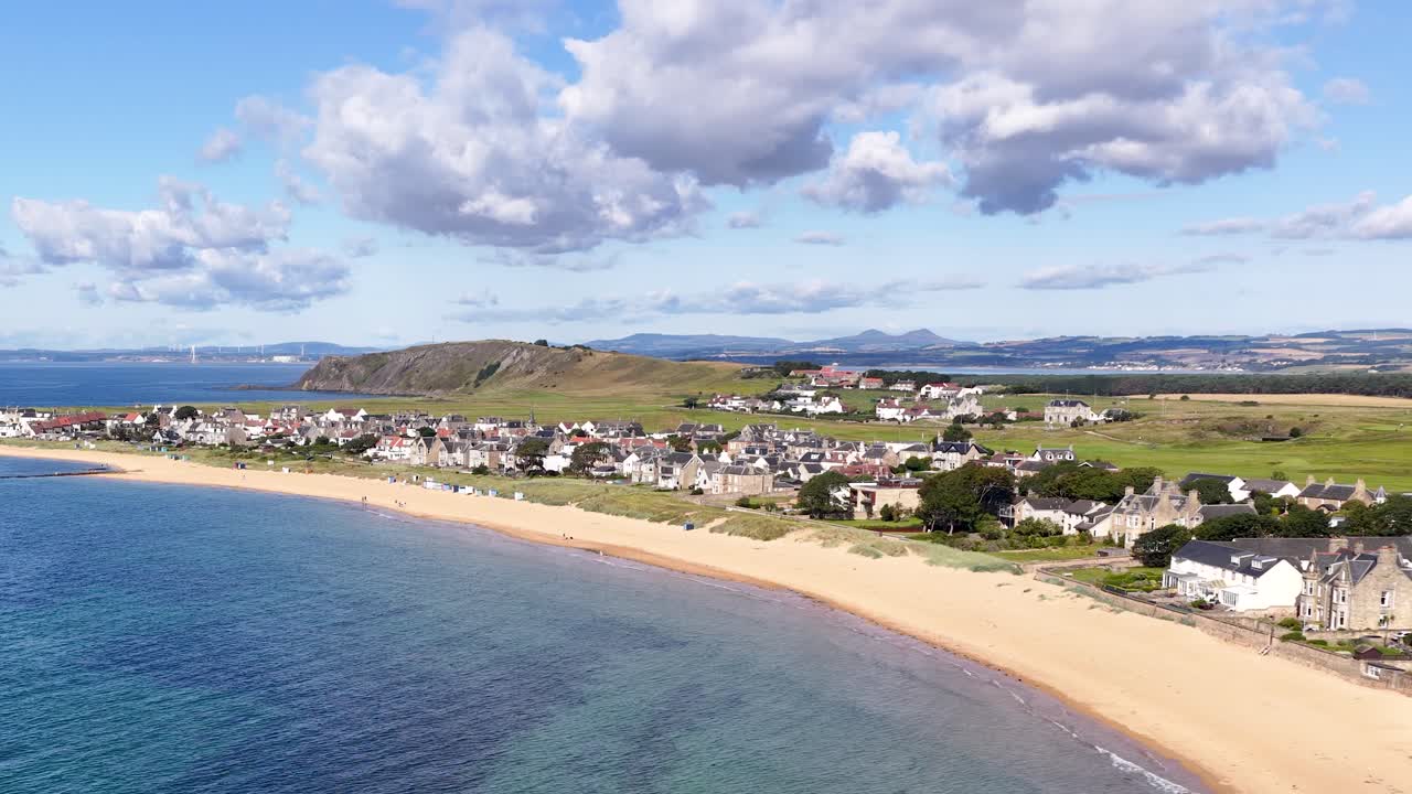 Drone glides over seaside village, sandy shoreline, blue sea, and rolling hills under daylight