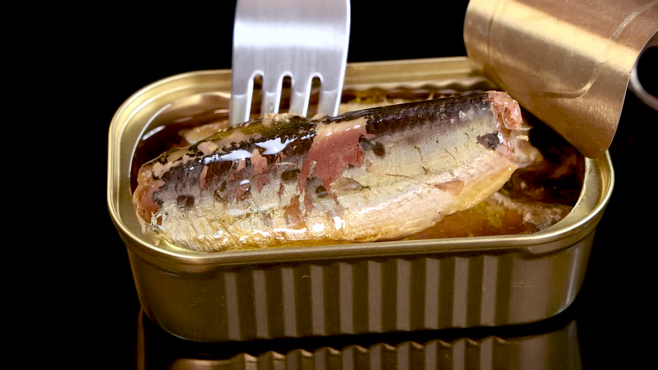 A metal fork lifts a sardine from an open tin can filled with oil, under bright studio lighting, with a black reflective background and steady camera angle