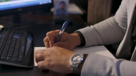 Mano-De-Hombre-De-Negocios-Sosteniendo-Un-Bolígrafo-Escribiendo-En-Papel-Frente-A-Su-Computadora