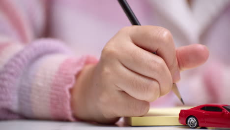 Close-up-of-a-hand-holding-a-pencil-and-writing-something-on-a-yellow-paper,-behind-a-red-toy-car-that-signifies-car-loan-financing