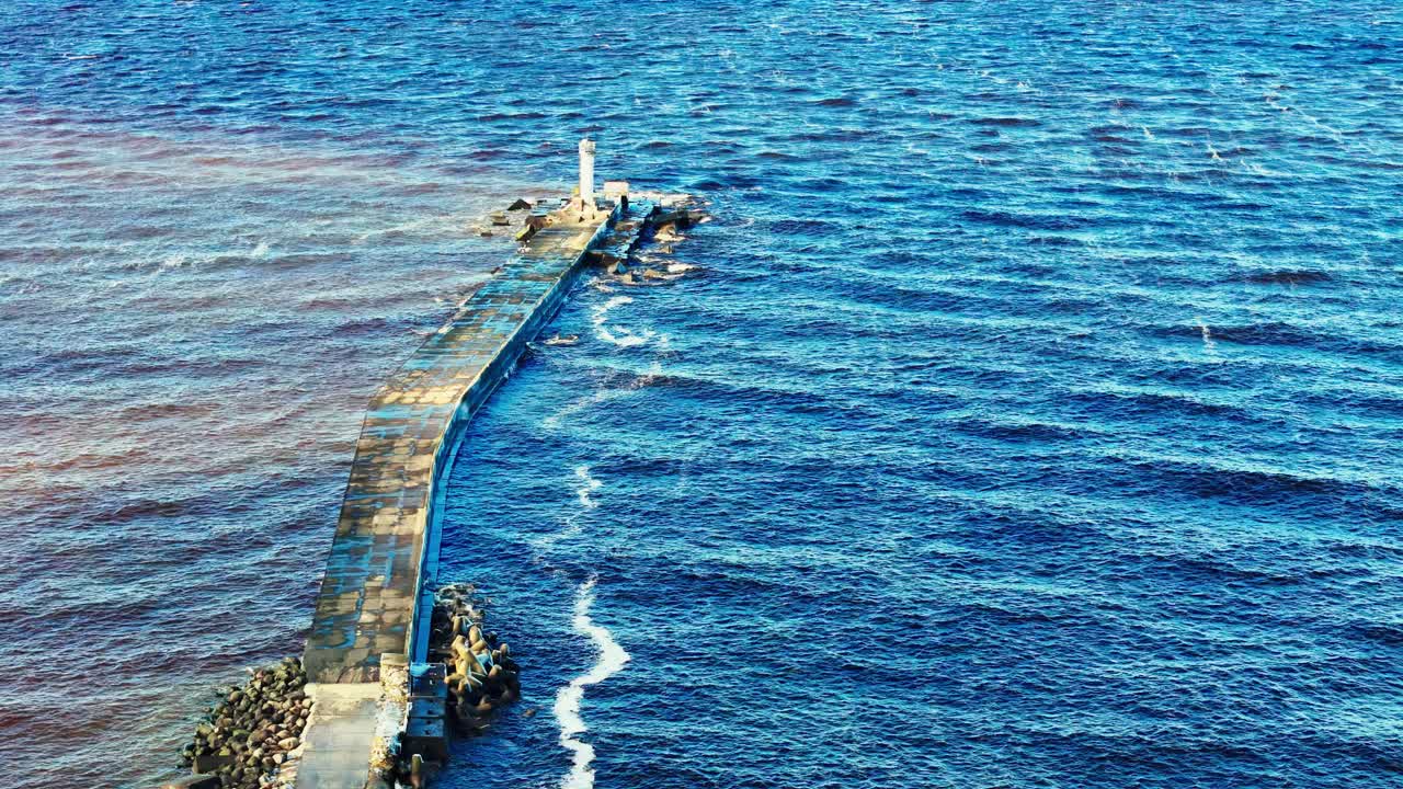 Bolderāja breakwater in Riga, Latvia, on a bright, sunny day, showing the distinct color difference between the dark blue river water and the lighter, grey-blue sea, with a small lighthouse