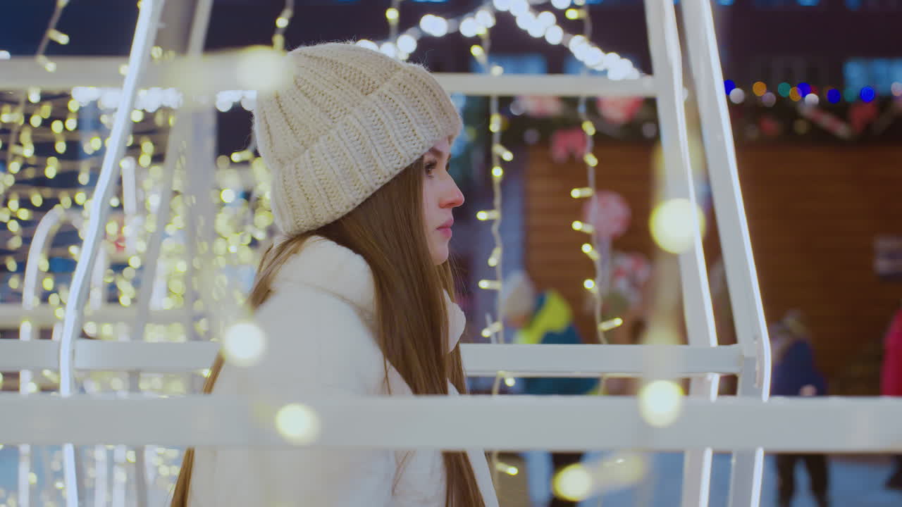 Elegant woman in white beanie and winter jacket walking past glowing golden holiday lights at night, festive atmosphere with decorative string lights illuminating surroundings