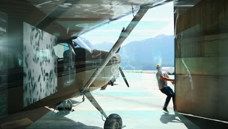 In hangar, man opening door of small aircraft with mountains in background