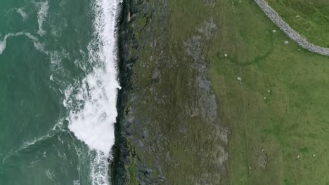 abstract topdown half and half aerial of green rocky coastal cliffs and rough turquoise sea waves running along the edge of the headland