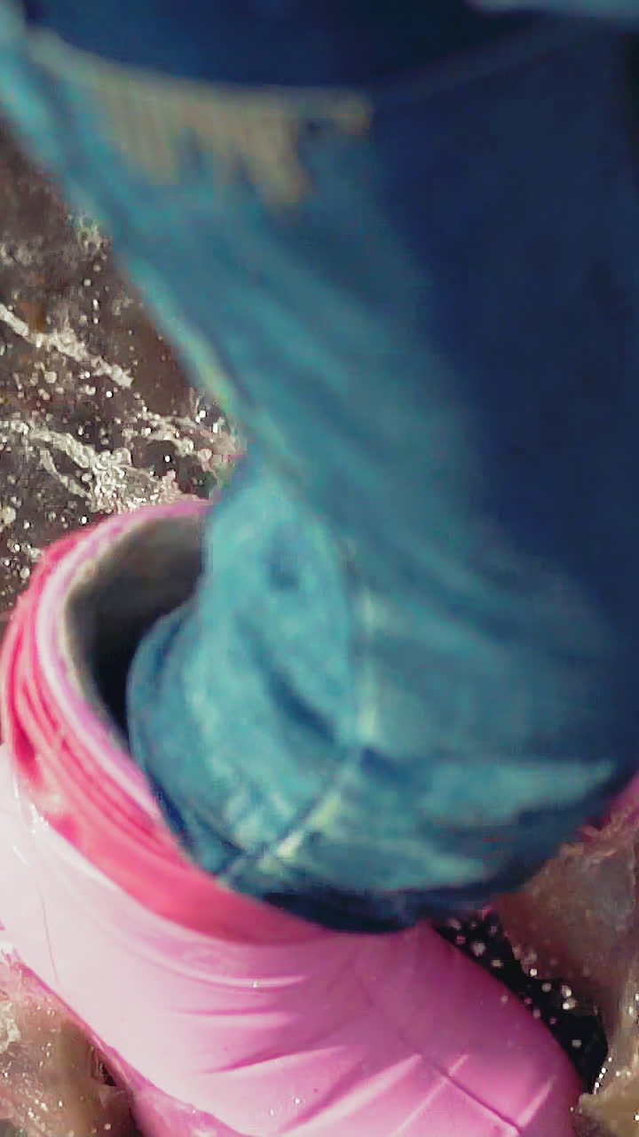 CU shot, slow motion: active little child wearing bright pink rubber boots and jeans has fun jumping in puddle on sunny day after rain extreme closeup