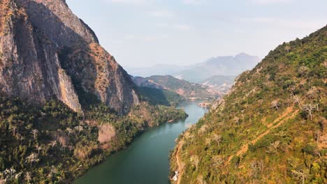slow aerial glide over the nam ou river as it carves through the dramatic limestone mountains of nong khiaw, northern laos, with visible jungle-clad cliffs and karst formations dominating the horizon